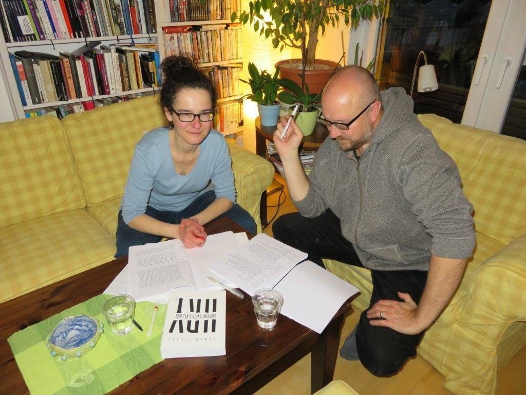 Barbora Schnelle und Roman Sikora auf gelben Sofas vor Blättern mit dem Stücktext und seiner Übersetzung. Im Hintergrund eine Bücherwand und Pflanzen.
